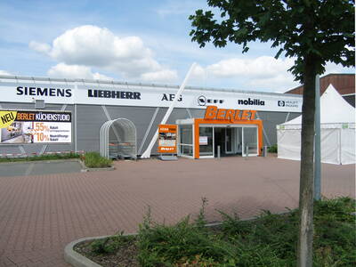 Eingangsbereich eines Berlet-Elektromarkts mit orangem Vordach, Werbeplakat für ein neues Küchenstudio und Einkaufswagenbereich auf einem gepflasterten Parkplatz unter bewölktem Himmel