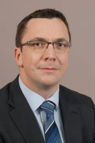 Mittelalterlicher Mann mit Brille im dunklen Anzug und blau gestreifter Krawatte blickt ernst in die Kamera vor neutralem Hintergrund