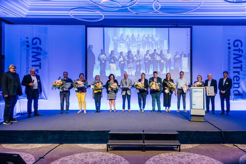 Auszeichnungsgala der GfMTrend mit einer Reihe von Preisträgern, die auf der Bühne Blumensträuße und Urkunden vor einer großen Leinwand mit ihrem Gruppenfoto präsentieren