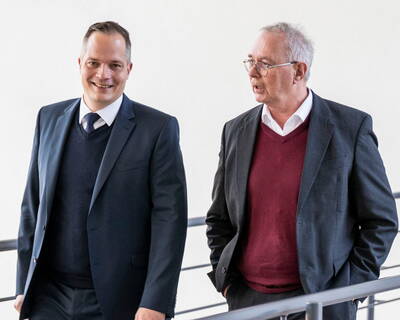 Zwei Geschäftsmänner im Anzug gehen nebeneinander einen Flur entlang und unterhalten sich lebhaft.
