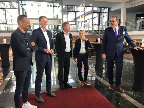 Fünf elegant gekleidete Geschäftsleute stehen lachend in einer modernen, lichtdurchfluteten Empfangshalle auf rotem Teppich und unterhalten sich locker miteinander.