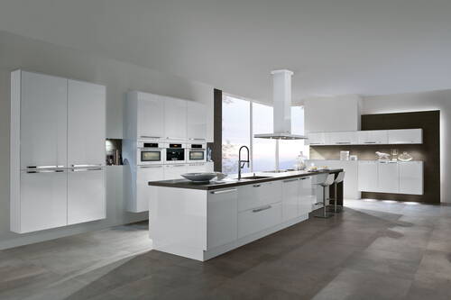 Helle, moderne Einbauküche mit weiß glänzenden Fronten, Kochinsel mit Spülbecken und Barhockern, deckenhohen Schränken und integrierten Elektrogeräten vor großen Fenstern.