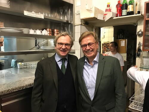 Zwei elegant gekleidete Männer lächeln nebeneinander in einer belebten Restaurantküche mit sichtbarer Theke, Geschirr und Personal im Hintergrund