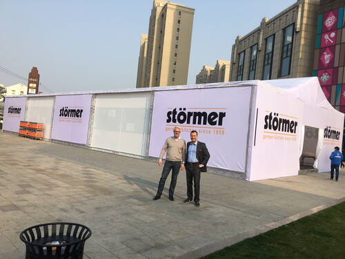 Zwei Männer posieren vor einem großen weißen Messezelt mit dem Aufdruck „Störmer – german kitchens since 1958“ auf einem modernen Platz mit hohen Gebäuden im Hintergrund.