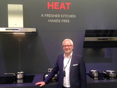 Lächelnder Mann im Anzug präsentiert moderne AEG-Dunstabzugshauben und Kochfelder vor einer dunkelgrauen Messewand mit dem Schriftzug „HEAT – A FRESHER KITCHEN HANDS FREE“.
