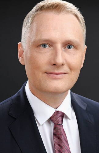 Blonder Mann im Anzug mit weißem Hemd und roter Krawatte blickt lächelnd direkt in die Kamera vor dunklem Hintergrund