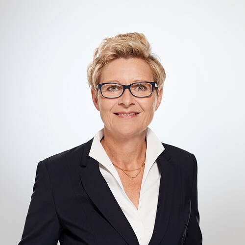 Lächelnde Geschäftsfrau mit kurzem blondem Haar und Brille in dunkelblauem Blazer vor hellem Hintergrund