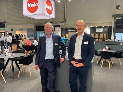 Zwei Geschäftsleute im Anzug stehen lächelnd am Messestand von Hailo in einer gut besuchten Ausstellungshalle