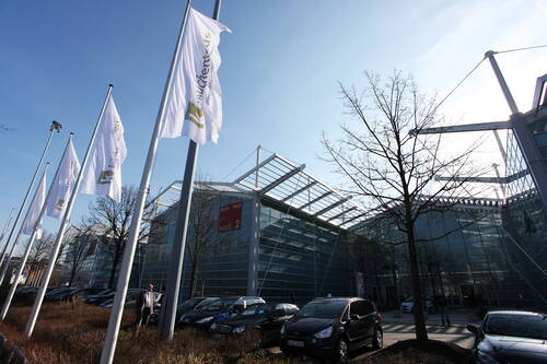 Moderne Messehalle mit verglaster Fassade, wehenden Messefahnen und geparkten Autos an einem sonnigen Tag