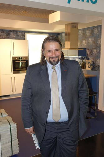 Mittelalterlicher Mann im grauen Anzug mit beigefarbener Krawatte lächelt in einer modernen Küchenausstellung und hält ein Buch in der Hand.