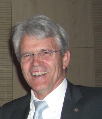 Älterer Mann mit grauen Haaren, Brille und Anzug lächelt breit in einem Innenraum mit holzvertäfelter Wand.