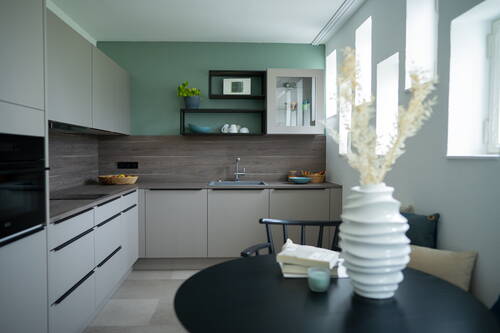 Helle, moderne Küche mit grauen Schränken, Holzarbeitsplatte, grüner Wand, offenem Regal über der Spüle und rundem Esstisch im Vordergrund mit weißer Vase und Büchern.