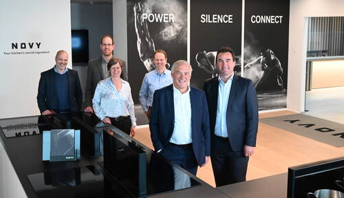Sechs geschäftlich gekleidete Personen stehen lächelnd in einem modernen Showroom von Novy vor einer Wand mit den Schlagworten „Power“, „Silence“ und „Connect“.