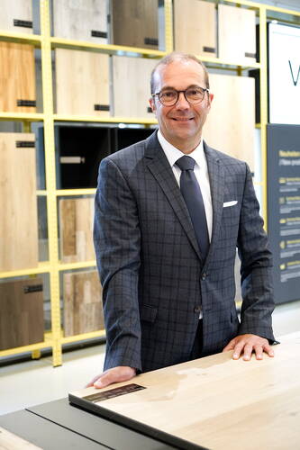Geschäftsmann im karierten Anzug steht lächelnd an einem Holztisch in einer modernen Ausstellung für Holz- und Möbeloberflächen