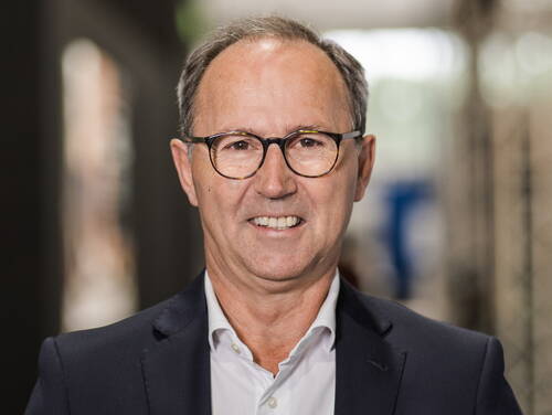 Älterer Mann mit Brille und Anzug lächelt freundlich in die Kamera vor unscharfem Hintergrund in Innenraumumgebung.