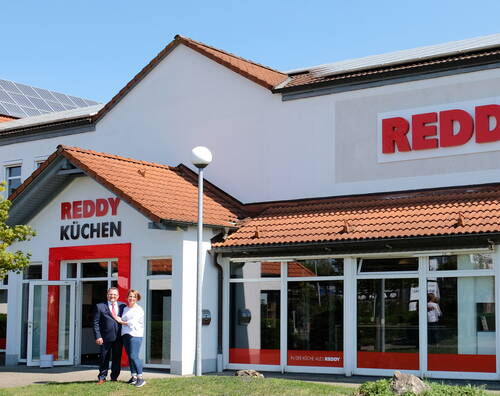 Zwei lächelnde Personen stehen vor dem Eingang eines Reddy Küchen Fachgeschäfts mit roten Dachziegeln und Solarpanelen auf dem Dach
