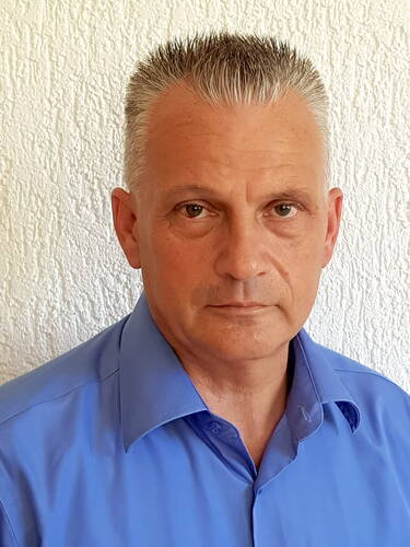 Mittelalterlicher Mann mit kurzem, graumeliertem Bürstenschnitt in einem blauen Hemd blickt ernst in die Kamera vor einer weißen Strukturwand