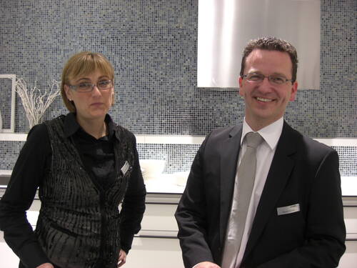 Zwei lächelnde Angestellte in Businesskleidung stehen vor einer modernen Wand mit Mosaikfliesen in einem hellen Innenraum