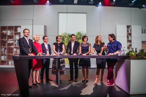 Neun elegant gekleidete Personen stehen an einer modernen Bar mit Sektgläsern, unterhalten sich ausgelassen und wirken wie bei einer festlichen Podiumsrunde in einem hell ausgeleuchteten Studio.