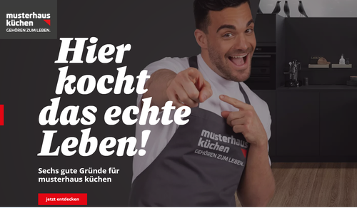 Lächelnder Koch in Schürze von „musterhaus küchen“ zeigt auf den Betrachter, daneben Werbeslogan „Hier kocht das echte Leben“ in moderner Küchenumgebung