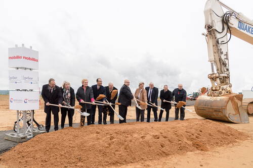 Politiker und Unternehmensvertreter stehen in einer Reihe auf einer sandigen Baustelle und heben beim symbolischen ersten Spatenstich mit Schaufeln Erde vor einem großen Bagger aus.
