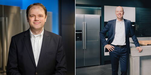 Zwei Geschäftsleute in dunklen Anzügen posieren in einer modernen Küche mit Edelstahl-Kühlschränken und Design-Arbeitsplatte.