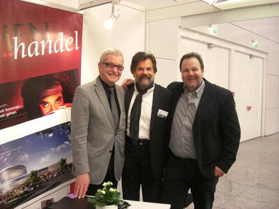 Drei lächelnde Männer in Anzügen stehen Arm in Arm vor einem Messestand mit Werbeplakaten und modernem Gebäudefoto in einer hellen Ausstellungshalle