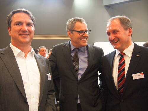 Drei geschäftlich gekleidete Männer stehen lachend in einem Konferenzraum zusammen und unterhalten sich in entspannter Atmosphäre.