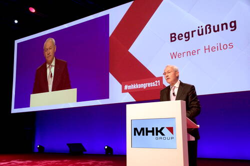 Redner am Pult der MHK Group begrüßt die Teilnehmer des MHK-Kongresses 2021 vor großer Leinwand mit seinem projizierten Bild und dem Schriftzug „Begrüßung – Werner Heilos“
