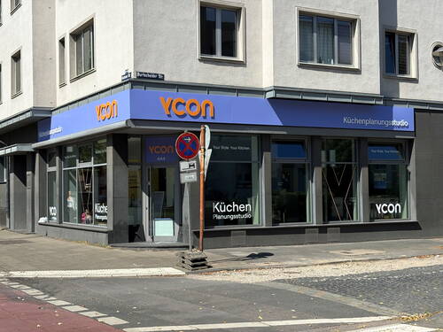 Modernes Eckgebäude mit blau-orangener VCON‑Beschilderung, das als Küchenplanungsstudio mit großen Schaufenstern an einer Straßenecke in der Sonne liegt