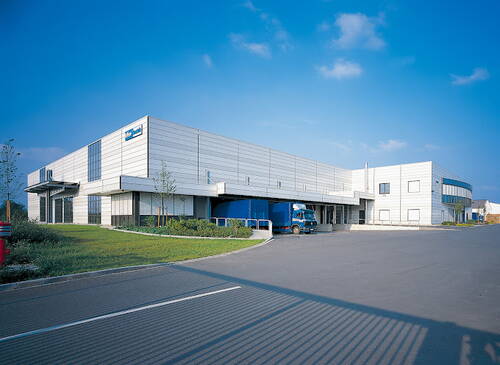 Moderner Logistik- und Industriekomplex mit Laderampen, an dem ein blauer Lkw unter klarem Himmel an einer weitläufigen asphaltierten Zufahrt steht.