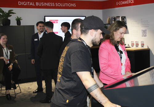 Messebesucher testen gemeinsam eine interaktive Touchscreen‑Anwendung an einem Stand für digitale Business‑ und E‑Commerce‑Lösungen, während im Hintergrund weitere Teilnehmende im Gespräch sind.