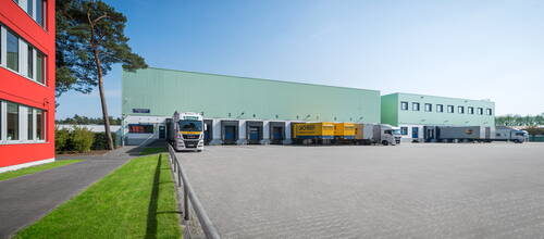 Mehrere Lastwagen be- und entladen an den Laderampen eines großen, mintgrünen Logistiklagers neben einem roten Bürogebäude an einem sonnigen Tag