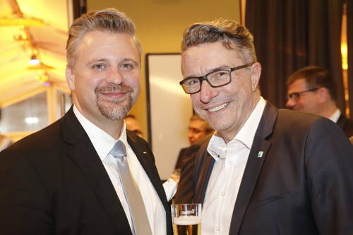 Zwei lächelnde Männer in Anzug und Krawatte stehen bei einem Empfang eng beieinander, einer hält ein Glas Bier in der Hand.