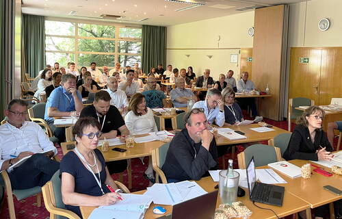 Teilnehmerinnen und Teilnehmer eines gut besuchten Seminars sitzen konzentriert in einem hellen Konferenzraum und machen sich Notizen während eines Vortrags.