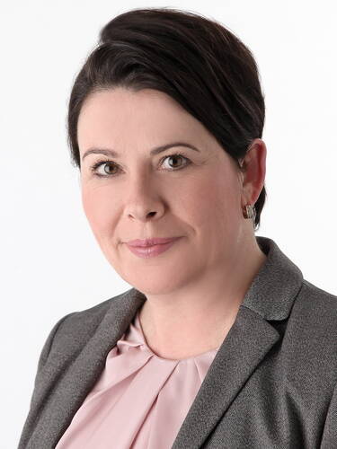 Frau mit kurz geschnittenem dunkelbraunem Haar im grauen Blazer und rosafarbenem Blusentop blickt freundlich in die Kamera vor weißem Hintergrund