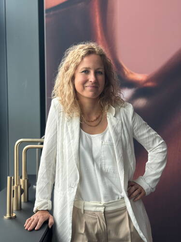 Lockenhaarige Frau in heller Bluse und weißem Blazer lehnt lächelnd an einer dunklen Arbeitsplatte mit goldenen Armaturen vor einer modernen, abstrakten Wandgestaltung