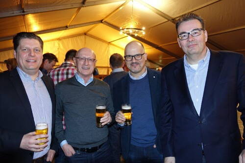 Vier Männer in Business-Kleidung stehen lächelnd in einem Festzelt dicht beieinander und halten jeweils ein Glas Bier in der Hand.