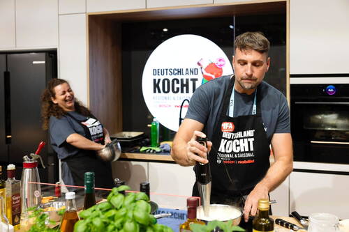 Mann püriert konzentriert eine cremige Sauce mit dem Stabmixer, während neben ihm eine lachende Frau in Schürze bei einem „Deutschland kocht“-Kochevent in einer modernen Showküche arbeitet.