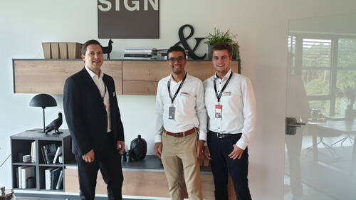 Drei lächelnde Geschäftsleute in weißem Hemd und Anzug stehen in einem modern eingerichteten Büro vor einem Holzregal mit Dekoobjekten und dem Schriftzug „SIGN“ an der Wand
