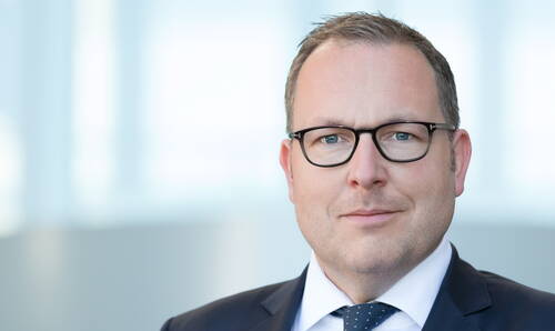 Geschäftsmann mit Brille und Anzug blickt ernst in die Kamera, vor unscharfem, hellen Bürohintergrund