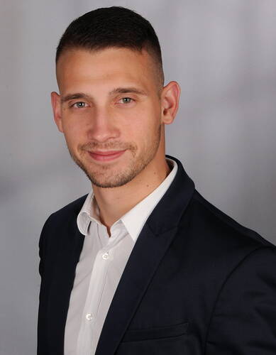 Junger Mann im Business-Anzug mit weißem Hemd lächelt freundlich in die Kamera vor neutral grauem Hintergrund