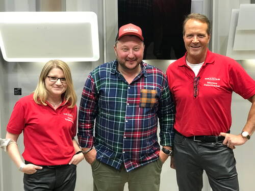 Drei lächelnde Personen posieren nebeneinander in einer modernen Ausstellungshalle, zwei in roten Firmenpoloshirts und eine in kariertem Hemd mit roter Kappe.