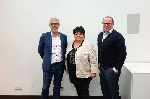 Drei lächelnde Geschäftsleute stehen nebeneinander vor einer weißen Wand in einem Konferenzraum.
