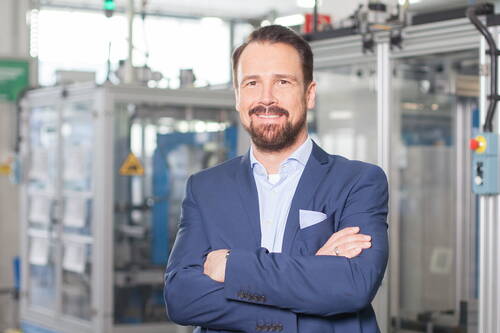 Geschäftsmann im blauen Anzug steht mit verschränkten Armen lächelnd in einer modernen Industriehalle mit Maschinen im Hintergrund