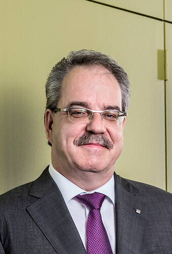 Mittelalterlicher Mann mit Brille, grauem Haar und Schnurrbart in dunklem Anzug und lila Krawatte lächelt leicht vor einer beigefarbenen Wand.