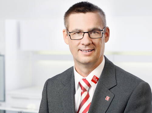 Mittelalterlicher Mann mit Brille im grauen Anzug und rot‑weiß gestreifter Krawatte lächelt vor einem hellen, modernen Bürohintergrund in die Kamera.