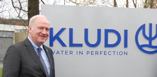 Älterer Mann im Anzug lächelt vor einem großen Firmenschild mit dem blauen Schriftzug „KLUDI – WATER IN PERFECTION“ im Außenbereich.