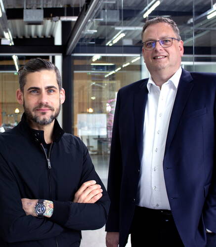 Zwei Männer in Businesskleidung stehen nebeneinander in einem modernen Büro mit Glaswänden und schauen lächelnd in die Kamera.