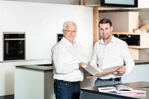 Zwei Männer in weißer Geschäftskleidung stehen in einer modernen Ausstellungsküche und vergleichen gemeinsam Musterplatten für Arbeitsflächen.